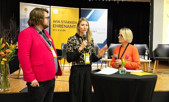 Drei Personen stehen an Rednertisch  und sprechen vor den Gästen