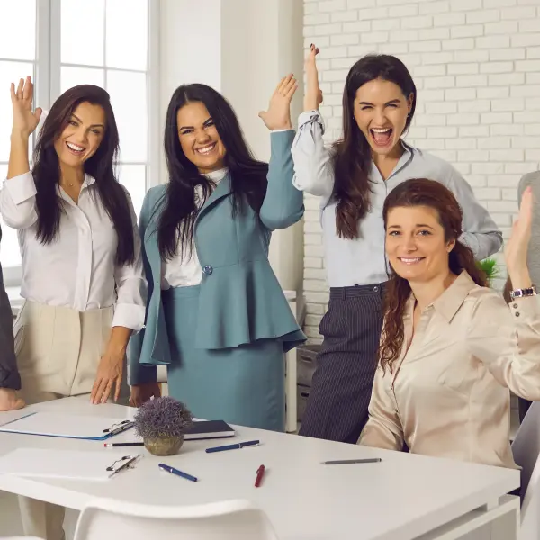 Sechs Frauen reihen sich um einen Schreibtisch und heben fröhlich die Hand in die Höhe