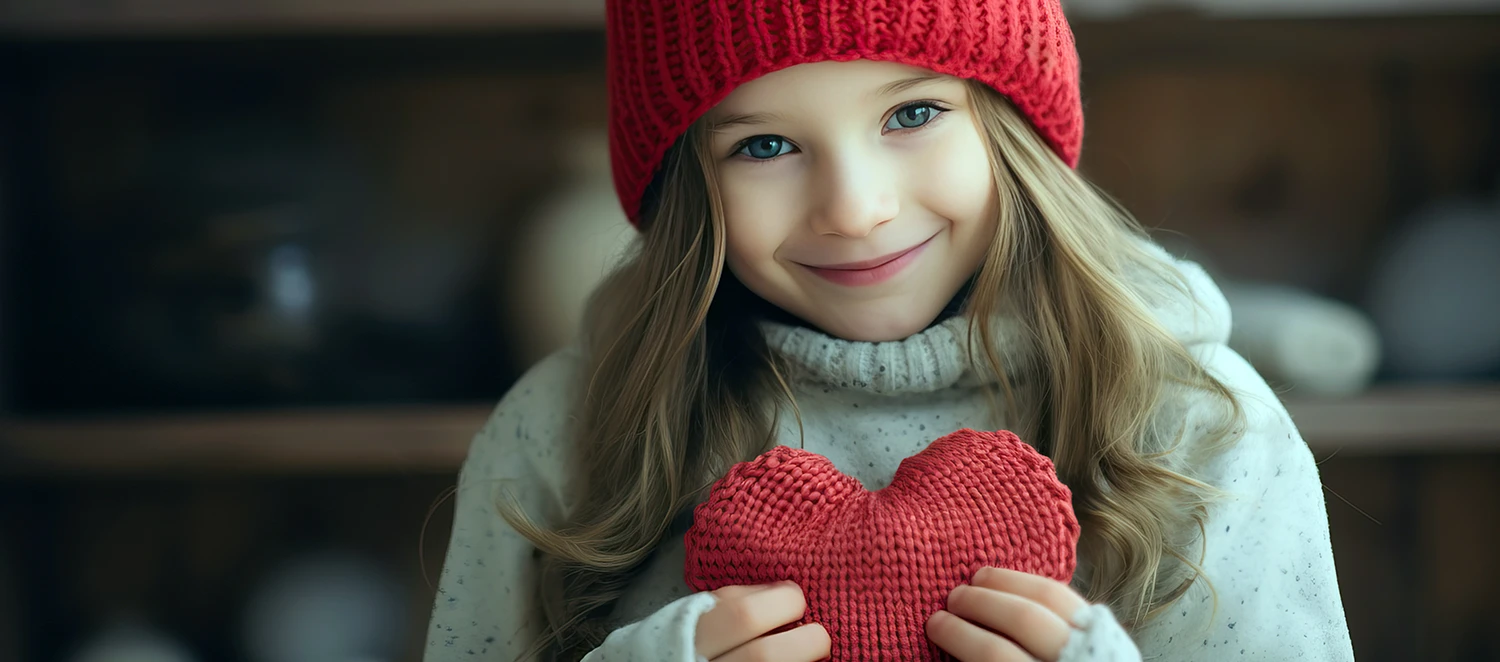 Ein kleines Mädchen im Pullover und roter Mütze lächelt und hält ein gestricktes Herz in ihren Händen. 